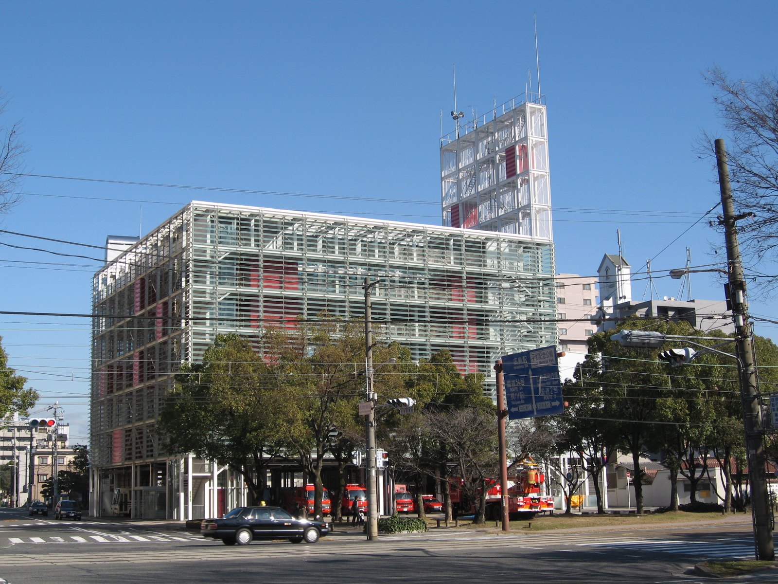 廣島市西消防署的玻璃格柵立面，隱約可見內部紅色消防器材，地面層停著消防車，右後方為訓練塔
