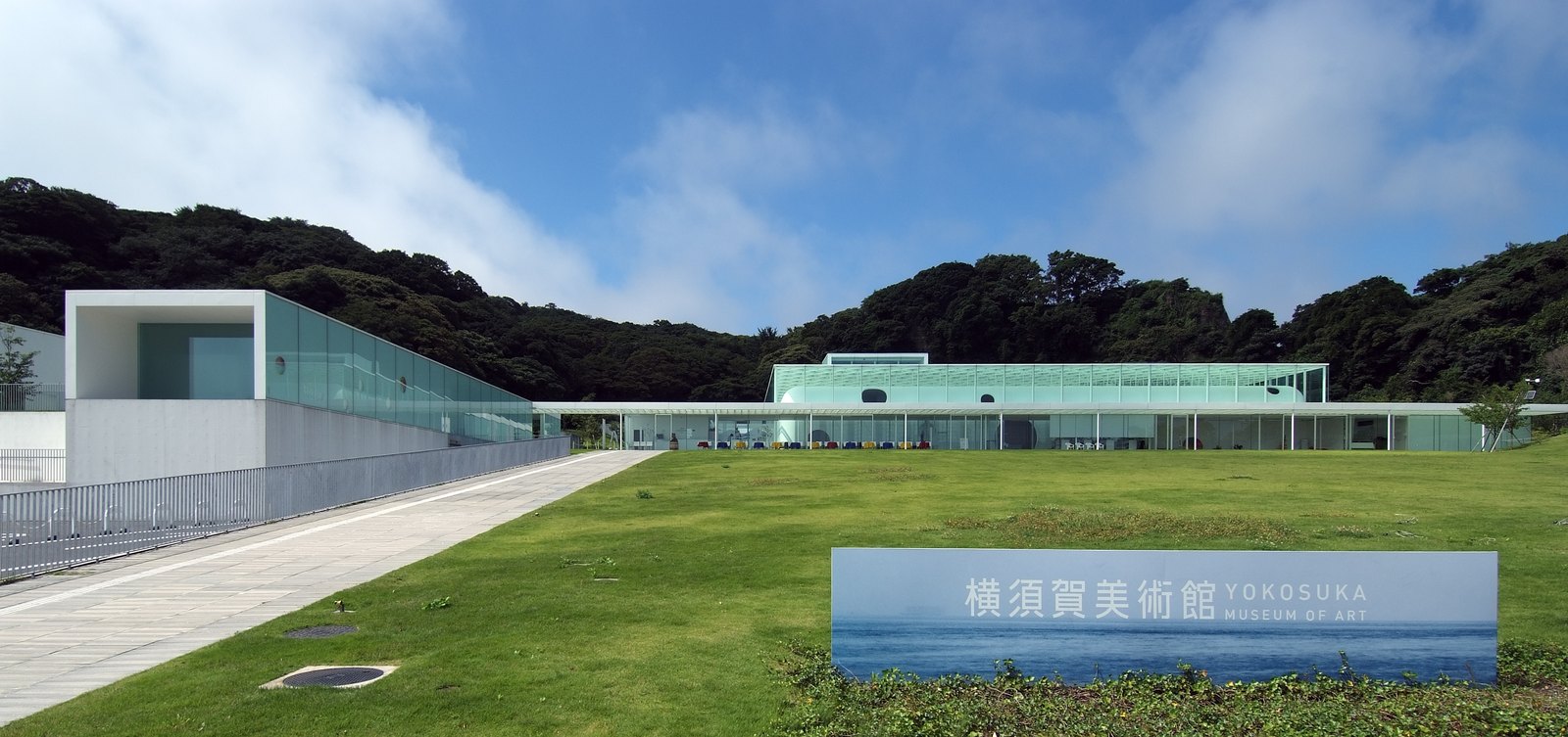 橫須賀美術館外觀，草地上的低矮量體，玻璃外皮下的白色鋼板上開著圓孔，背後為觀音崎的樹林